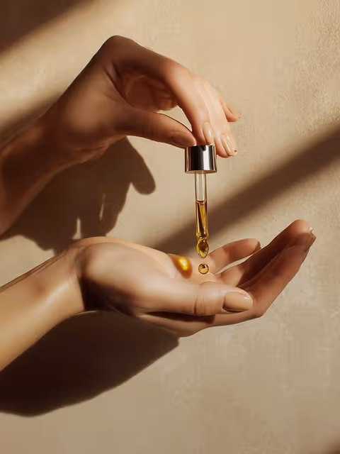 Close-up of two hands with one holding a dropper releasing golden oil droplets onto the other palm.