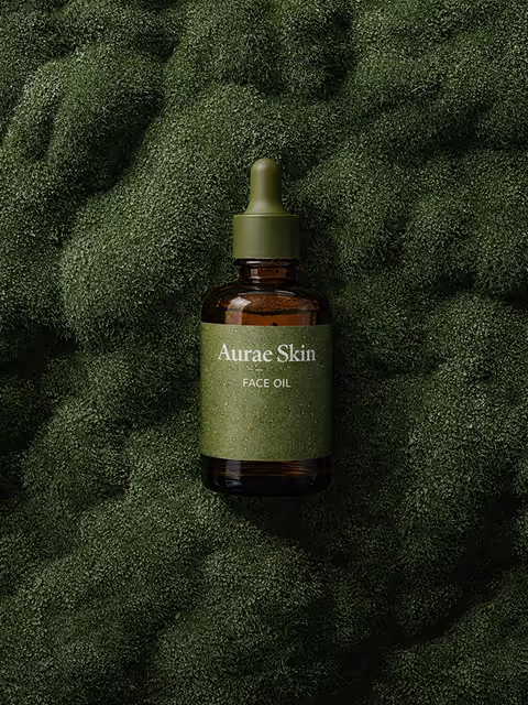 Amber glass dropper bottle labeled 'Aurae Skin Face Oil' resting on textured green moss.
