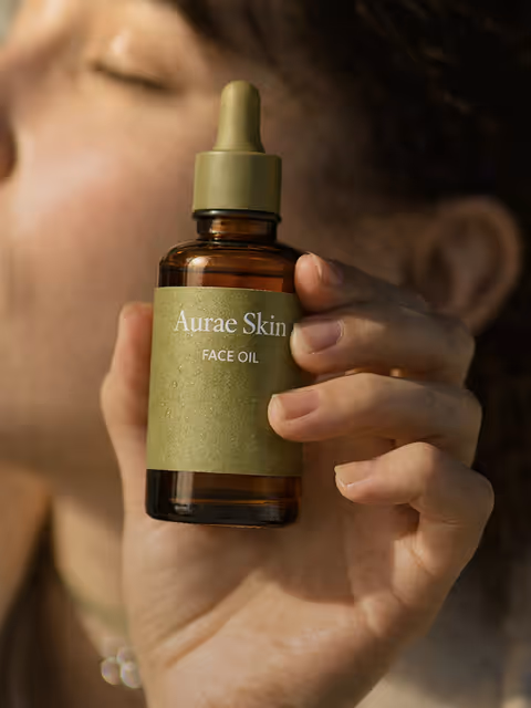 Close-up of a hand holding a brown glass dropper bottle labeled 'Aurae Skin Face Oil' with a blurred person in the background.